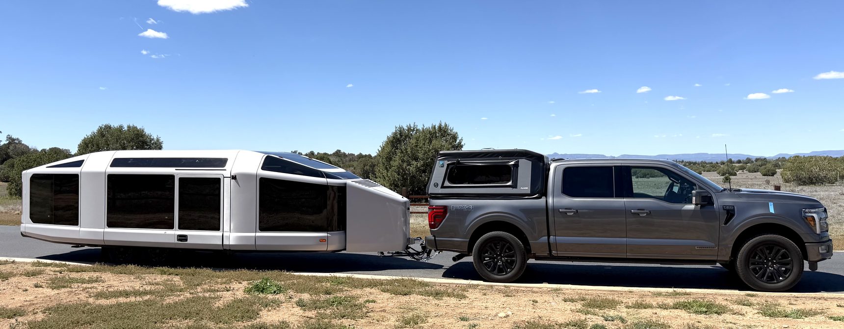 Ford F-150 PowerBoost in silver, with a bed topper, hitched to a silver LightShip AE.1 travel trailer on a paved pullout under a clear sky, high-desert scrub and distant mesas behind